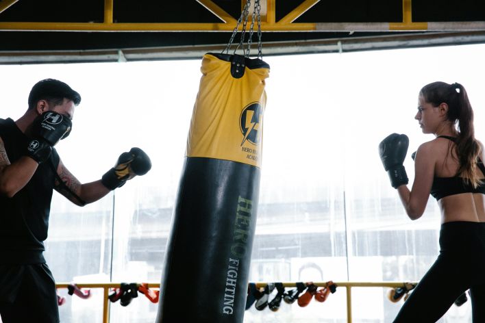 Two people engage in boxing training with a punching bag indoors, focusing on technique and strength.