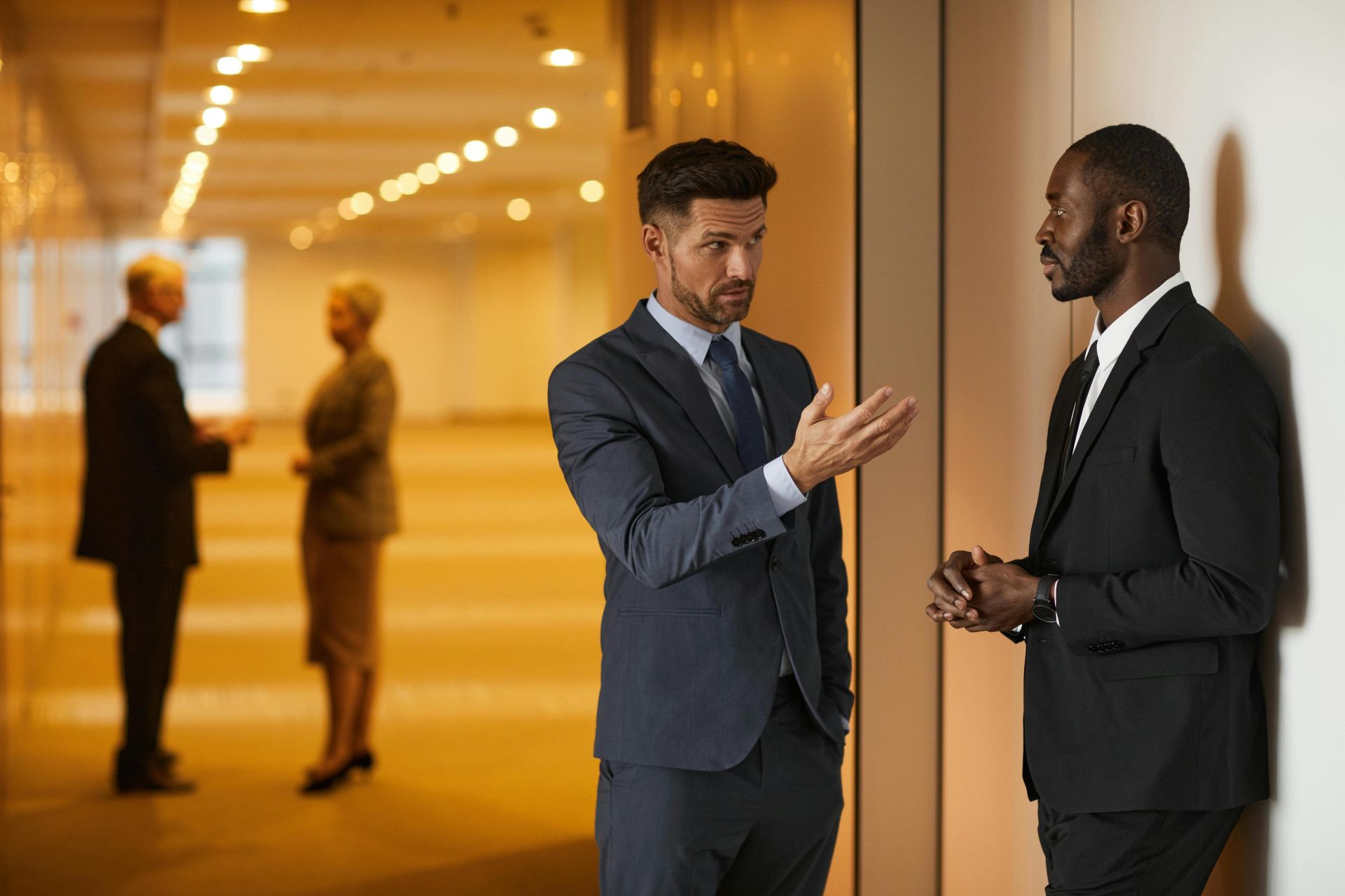 Two businessmen in formal attire engaged in a serious discussion in a modern office hallway.